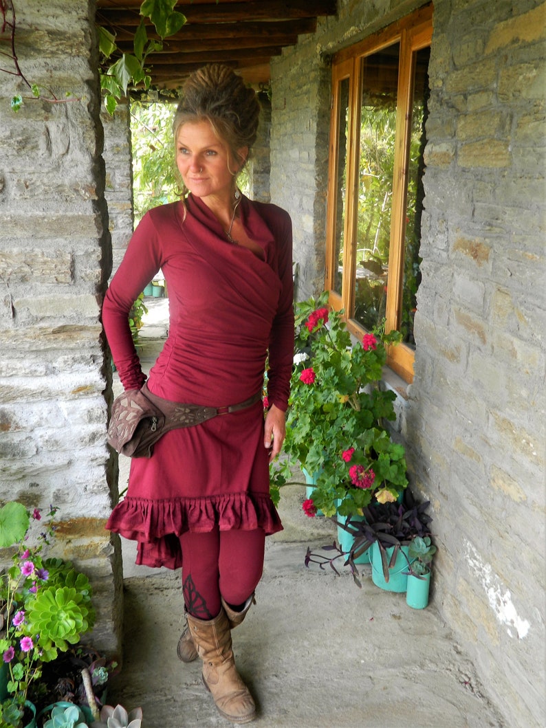 May include: A woman wearing a maroon wrap dress with a brown leather belt and brown boots. She is standing in a doorway with plants and stone walls.