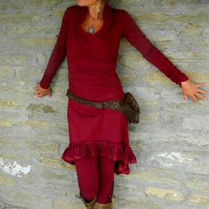 May include: A woman wearing a long-sleeved, burgundy dress with a ruffled hem. She is wearing a brown leather belt with a decorative buckle and a brown leather bag. She is standing in front of a stone wall.
