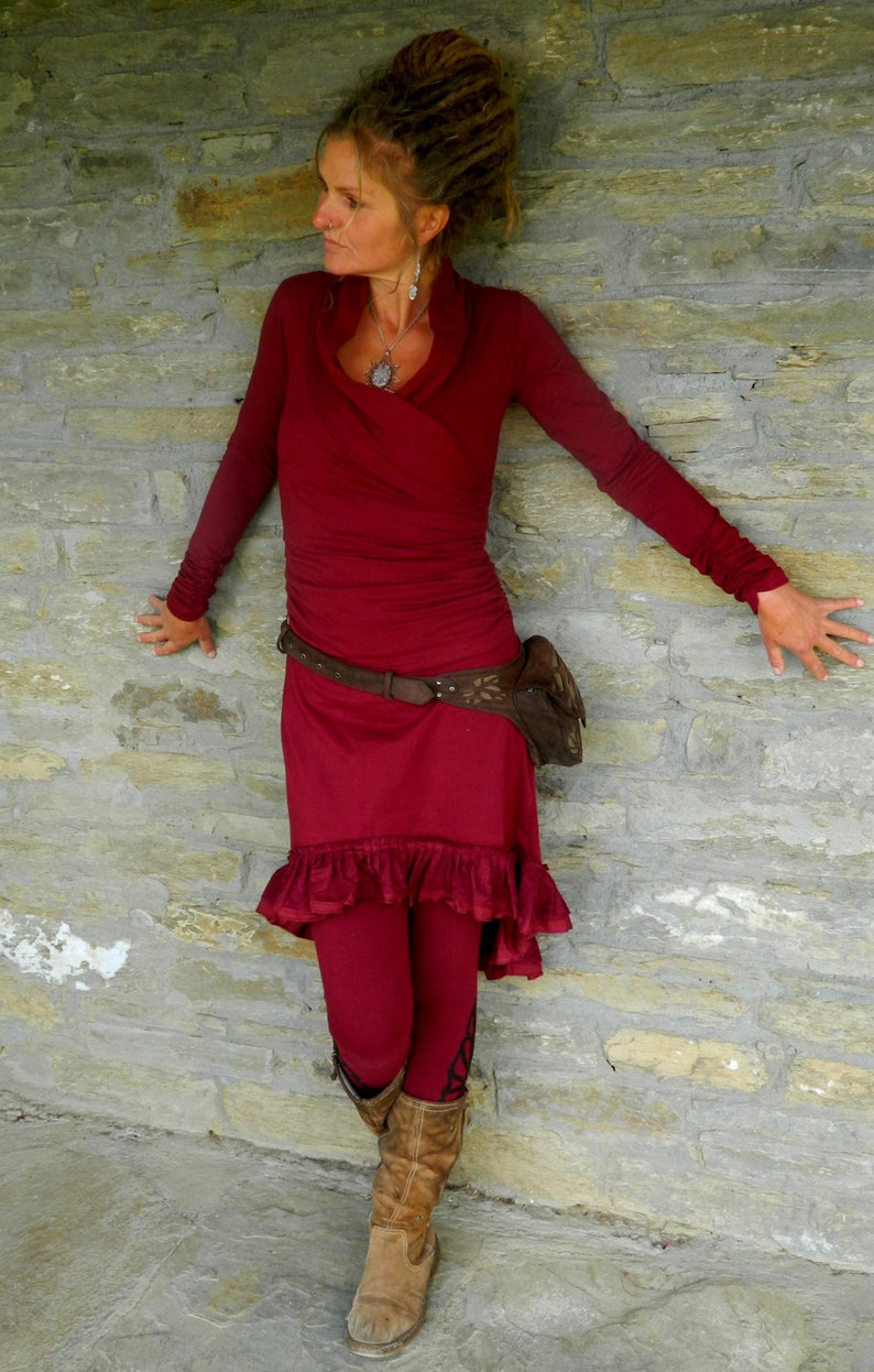 May include: A woman wearing a long-sleeved, burgundy dress with a ruffled hem. She is wearing a brown leather belt with a decorative buckle and a brown leather bag. She is standing in front of a stone wall.