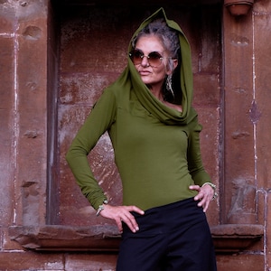 May include: A woman wearing a green long-sleeved shirt with a cowl neck and a hood. She is wearing sunglasses and has her hands on her hips.