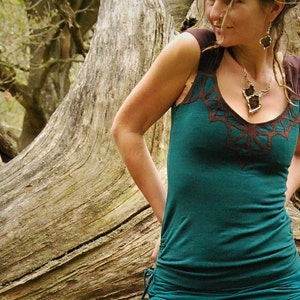 Può includere: Una donna indossa una canotta color teal con bordi marroni e un design geometrico. Indossa una collana d'argento con un ciondolo grande e orecchini d'argento.
