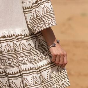 May include: A beige dress with flared sleeves and a tiered skirt, featuring a repeating geometric pattern in a dark brown color. A silver bracelet with dark accents adorns the wrist.
