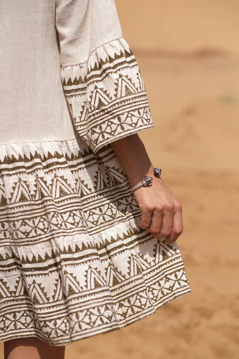 May include: A beige dress with flared sleeves and a tiered skirt, featuring a repeating geometric pattern in a dark brown color. A silver bracelet with dark accents adorns the wrist.
