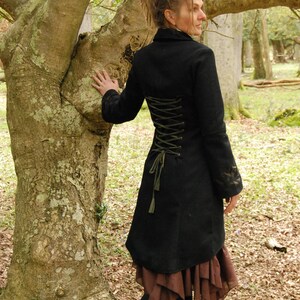 May include: A black wool coat with a long, fitted silhouette and a lace-up back detail. The coat has a high collar and long sleeves. The model is wearing the coat with a brown skirt and black boots.