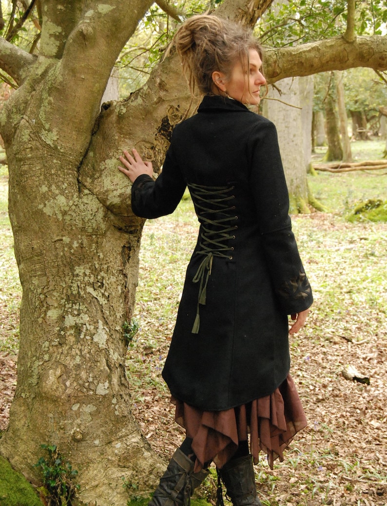 May include: A black wool coat with a long, fitted silhouette and a lace-up back detail. The coat has a high collar and long sleeves. The model is wearing the coat with a brown skirt and black boots.