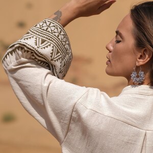 May include: A cream-colored linen shirt with intricate olive green and white patterned cuffs. The image also features silver floral earrings. The background is a blurred desert landscape.