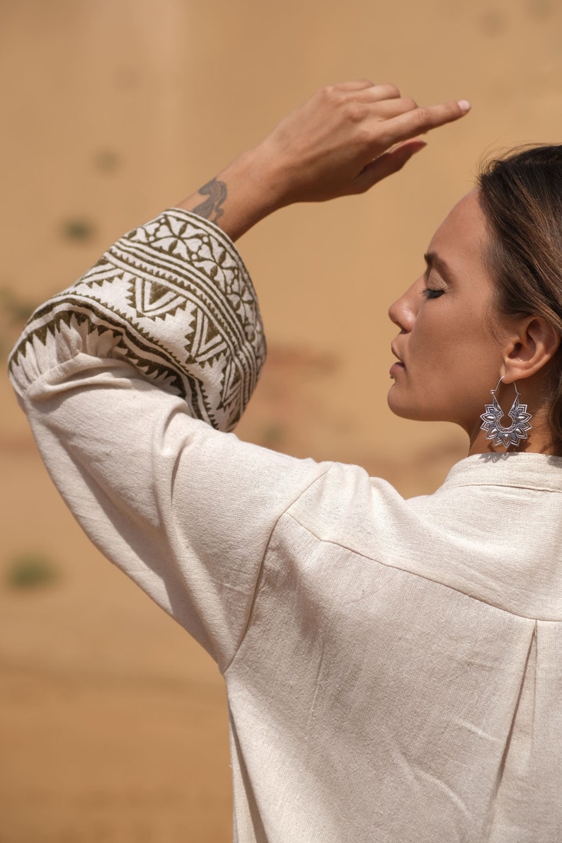 May include: A cream-colored linen shirt with intricate olive green and white patterned cuffs. The image also features silver floral earrings. The background is a blurred desert landscape.