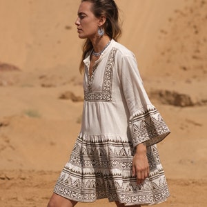 May include: A cream-colored, short-sleeved dress with brown geometric embroidery. The dress features a tiered skirt and flared sleeves, with the embroidery detailing the neckline, sleeves, and hem. The dress is worn outdoors.