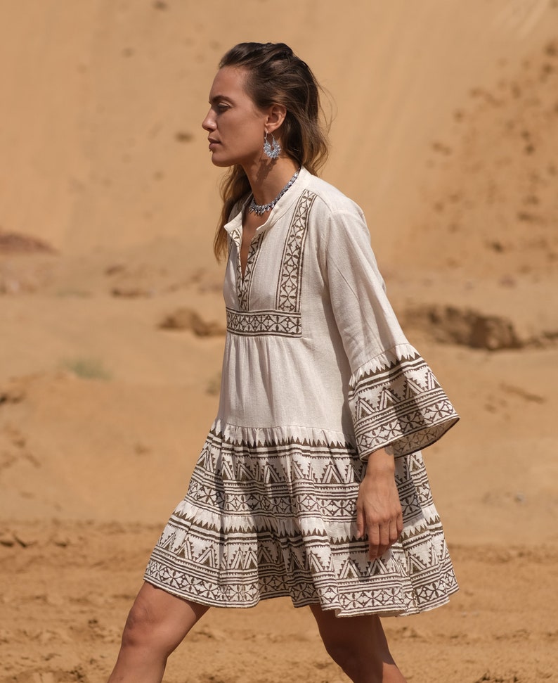 May include: A cream-colored, short-sleeved dress with brown geometric embroidery. The dress features a tiered skirt and flared sleeves, with the embroidery detailing the neckline, sleeves, and hem. The dress is worn outdoors.