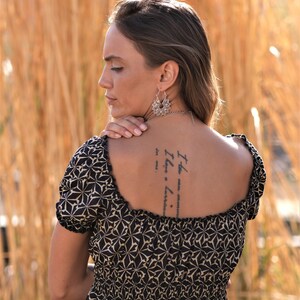 May include: A woman wearing a black and beige patterned dress with a square neckline. She has a tattoo on her back that reads "It the now... I have a... love... to me..."