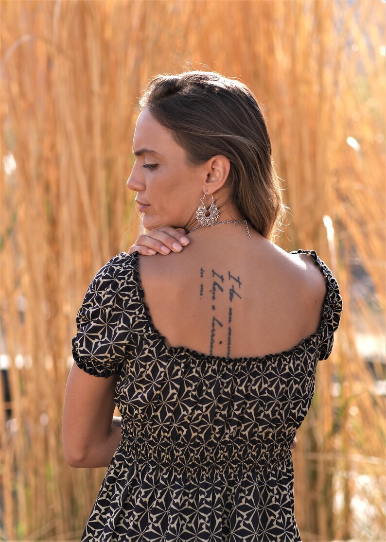 May include: A woman wearing a black and beige patterned dress with a square neckline. She has a tattoo on her back that reads "It the now... I have a... love... to me..."