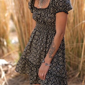 May include: A black and white patterned dress with a square neckline and short sleeves. The dress has a gathered waist and a tiered skirt. The model is wearing a silver necklace and bracelet.