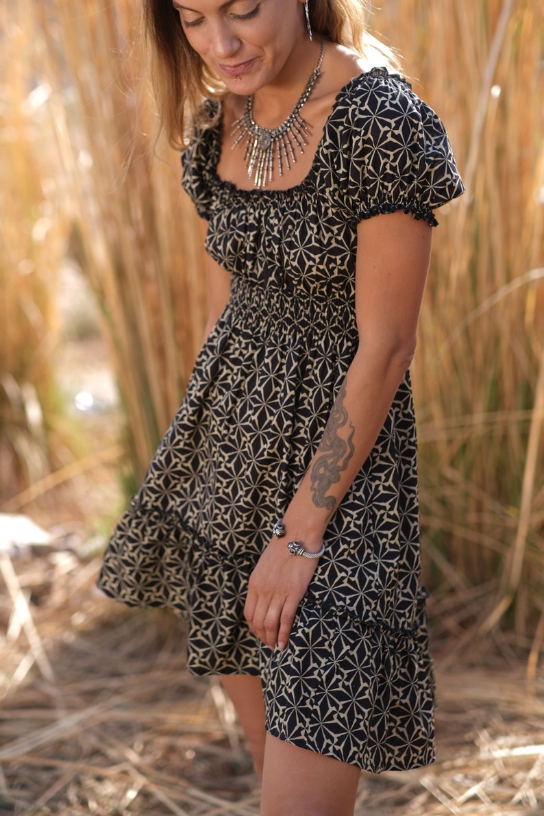 May include: A black and white patterned dress with a square neckline and short sleeves. The dress has a gathered waist and a tiered skirt. The model is wearing a silver necklace and bracelet.
