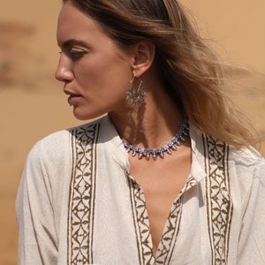 May include: A woman wearing a cream-colored tunic with a brown floral pattern, silver earrings, and a matching necklace. The earrings and necklace have a detailed, ornate design. The background is a blurred, sandy color.