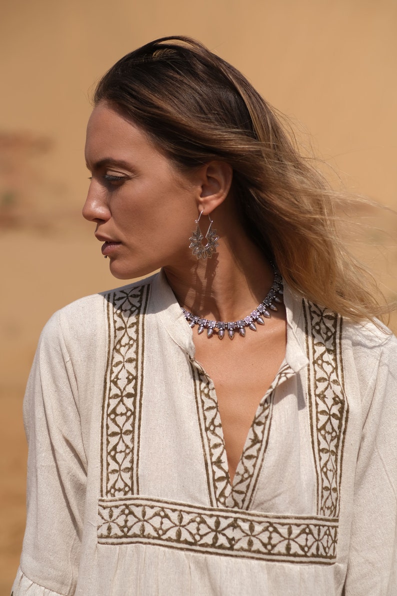 May include: A woman wearing a cream-colored tunic with a brown floral pattern, silver earrings, and a matching necklace. The earrings and necklace have a detailed, ornate design. The background is a blurred, sandy color.