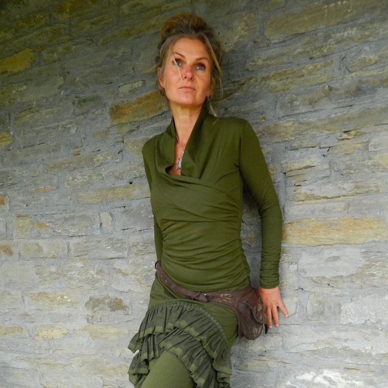 May include: A woman wearing a long-sleeved, olive green, wrap-style top with a ruffled skirt. She is wearing a brown belt with a decorative buckle and a brown leather fanny pack.