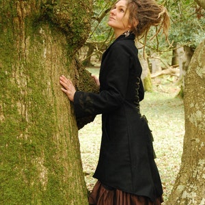 May include: A woman with long dreadlocks wears a black coat with a brown skirt and stands next to a moss-covered tree.