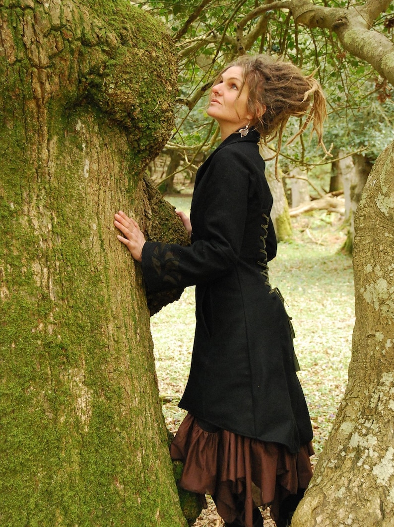 May include: A woman with long dreadlocks wears a black coat with a brown skirt and stands next to a moss-covered tree.
