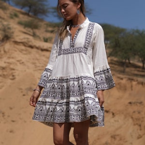 Puede incluir: Un vestido de algodón blanco con un patrón geométrico negro. El vestido tiene un escote en V, mangas largas y una falda de capas.