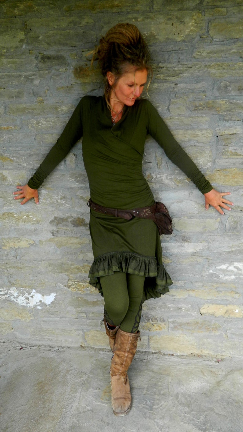 May include: A woman wearing a green long-sleeved shirt, a brown belt, and a green skirt with a ruffle hem. She is wearing brown boots and has her hair in dreadlocks.
