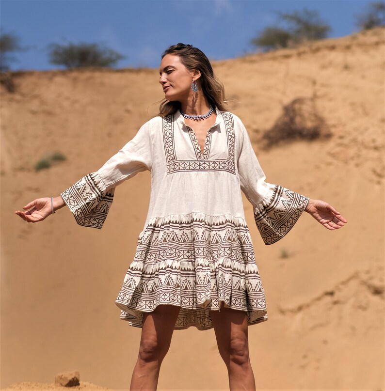 May include: A cream-colored, long-sleeved dress with brown geometric patterns on the sleeves and hem. The dress has a V-neck and is knee-length. The model is standing in a desert landscape with arms outstretched.
