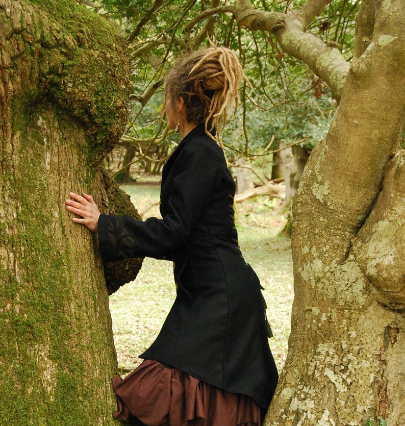 May include: A woman with long, blonde dreadlocks wears a black jacket with a brown skirt and stands next to a large tree trunk. The tree trunk is covered in moss and has a rough texture.