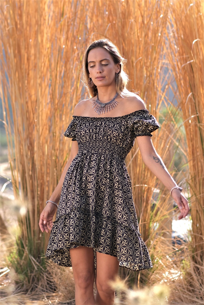 May include: A woman wearing a black and gold off-the-shoulder dress with a floral pattern. She is standing in front of a field of tall, dry grass.