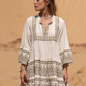 May include: A cream-colored, short dress with olive green geometric patterns. The dress features a V-neck, three-quarter sleeves, and a tiered skirt. The model is wearing a silver necklace and earrings.