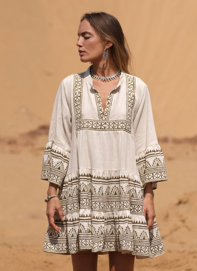 May include: A cream-colored, short dress with olive green geometric patterns. The dress features a V-neck, three-quarter sleeves, and a tiered skirt. The model is wearing a silver necklace and earrings.