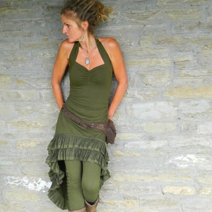 Puede incluir: Una mujer con un top verde de tirantes, un cinturón marrón y una falda verde con volantes. Está de pie frente a una pared de piedra.