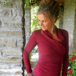 May include: A woman with dreadlocks wears a maroon long-sleeved shirt with a cowl neck. The shirt is made of a soft, stretchy fabric and has a relaxed fit.