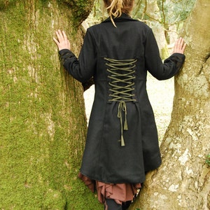 May include: A black coat with a long, flowing skirt and a green lace-up back. The coat is worn by a person with dreadlocks standing between two trees.