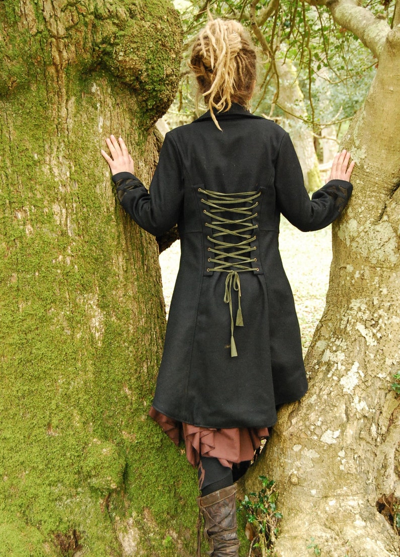 May include: A black coat with a long, flowing skirt and a green lace-up back. The coat is worn by a person with dreadlocks standing between two trees.