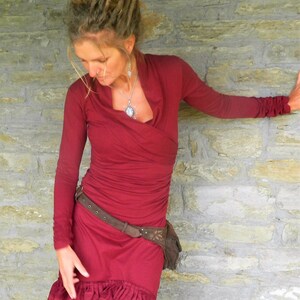 May include: A woman wearing a long-sleeved, burgundy dress with a ruffled hem. She is wearing a brown belt with a leather pouch attached. The dress has a V-neckline and is made of a soft, flowing fabric.