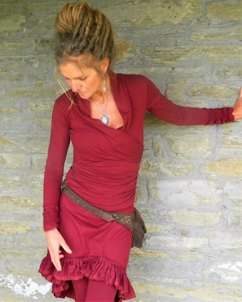 May include: A woman wearing a long-sleeved, burgundy dress with a ruffled hem. She is wearing a brown belt with a leather pouch attached. The dress has a V-neckline and is made of a soft, flowing fabric.