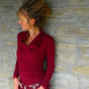 May include: A woman wearing a maroon long-sleeved wrap top with a deep V-neckline. She is standing in front of a stone wall.