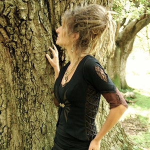 May include: A woman with dreadlocks is wearing a black lace top with brown accents. The top features a lace-up detail on the side and a spiderweb design on the front. She is standing next to a tree.