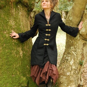 May include: A woman wearing a black jacket with gold buttons and a brown skirt stands in a forest. She is leaning against a tree trunk.