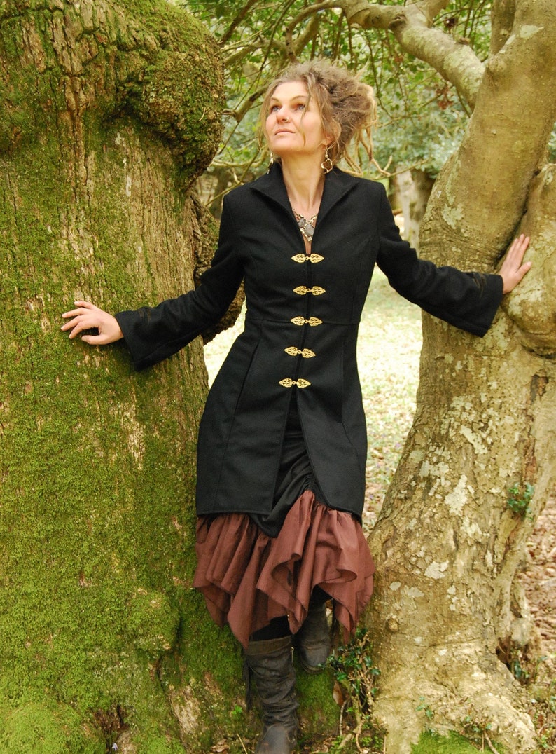 May include: A woman wearing a black jacket with gold buttons and a brown skirt stands in a forest. She is leaning against a tree trunk.