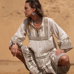 May include: A cream-colored dress with long sleeves and a V-neck, featuring geometric brown patterns on the bodice, sleeves, and skirt. The dress is paired with silver jewelry, including a necklace, earrings, and a bracelet. The model is squatting on sand.