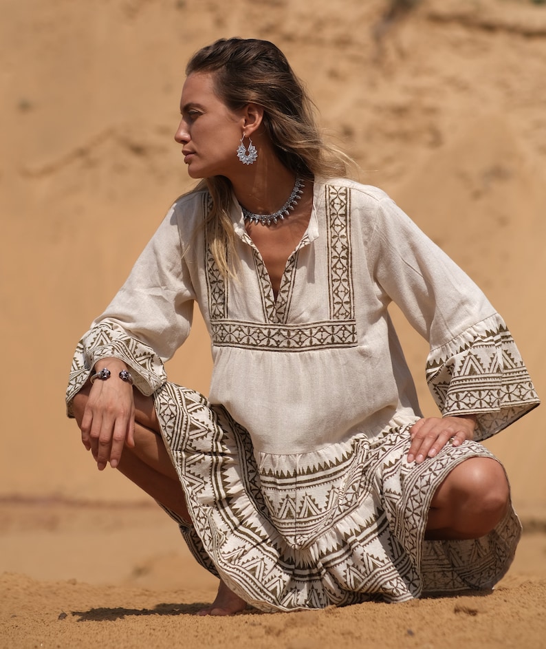 May include: A cream-colored dress with long sleeves and a V-neck, featuring geometric brown patterns on the bodice, sleeves, and skirt. The dress is paired with silver jewelry, including a necklace, earrings, and a bracelet. The model is squatting on sand.