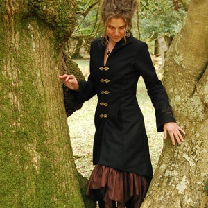 May include: A woman wearing a black coat with gold buttons and a brown skirt stands between two trees. She has long, brown hair with dreadlocks.
