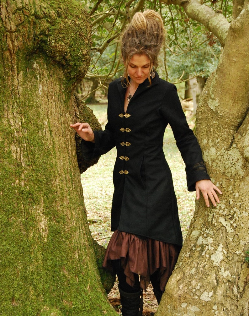 May include: A woman wearing a black coat with gold buttons and a brown skirt stands between two trees. She has long, brown hair with dreadlocks.