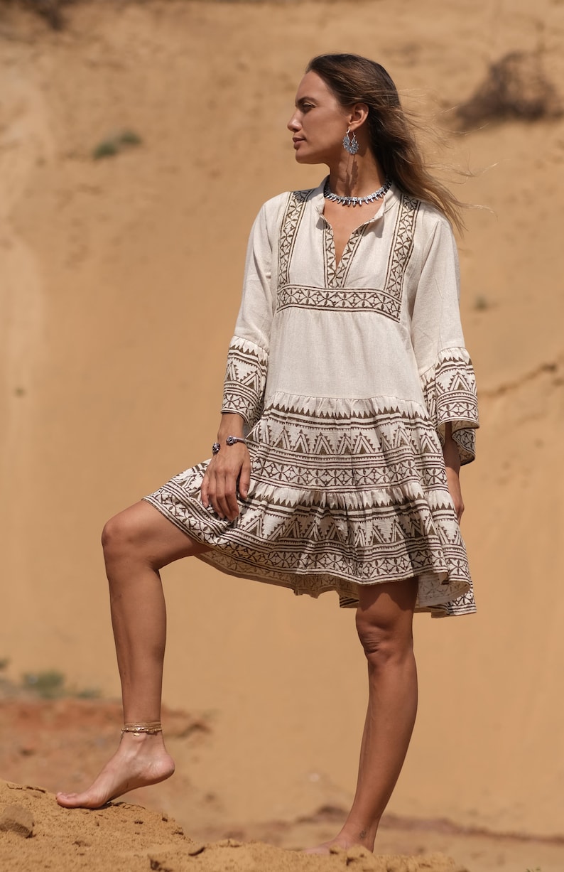 May include: A cream-colored, short-sleeved dress with brown geometric embroidery. The dress features a V-neck and a tiered skirt. The model is wearing silver earrings, a necklace, and a bracelet. The background is a sandy landscape.