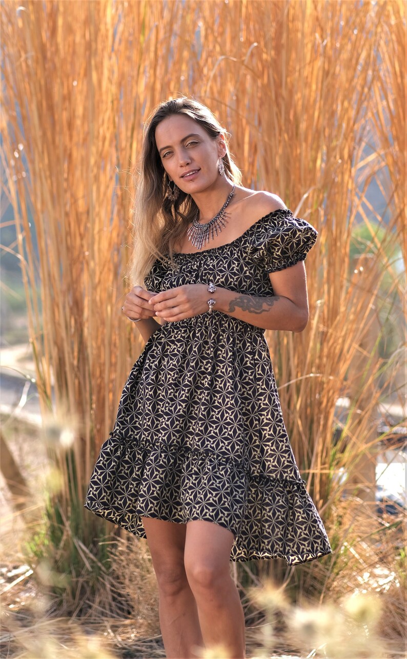 May include: A woman wearing a black and white floral off-the-shoulder dress with short puff sleeves. She is standing in front of a field of tall, dry grass.