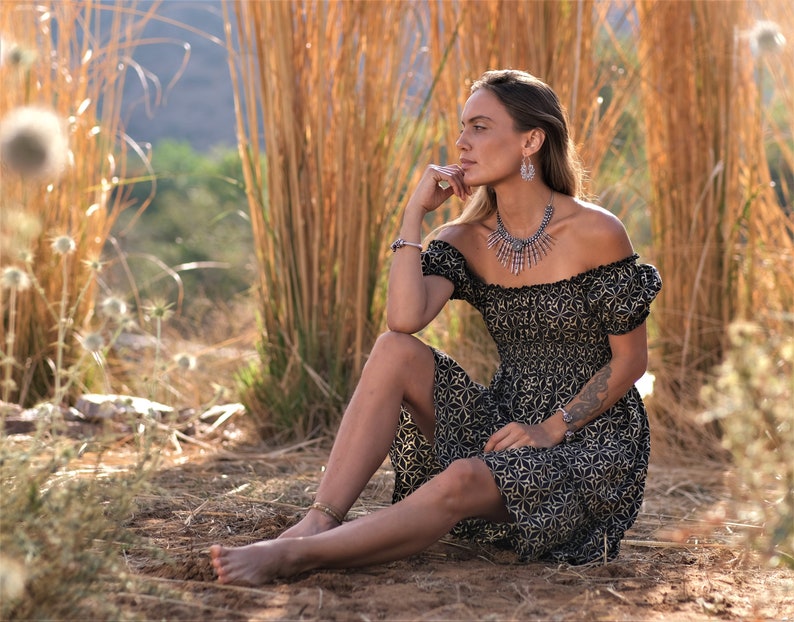 May include: A woman wearing a black and gold off-the-shoulder dress with a floral pattern sits in a field of tall grass.