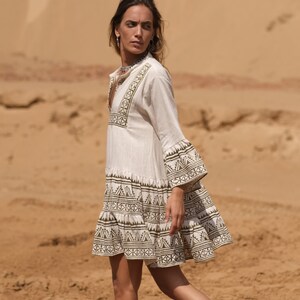 May include: A cream-colored, short dress with long sleeves and a flared skirt. The dress features intricate geometric patterns in a dark olive green color around the neckline, sleeves, and hem. The dress is made of a lightweight fabric, perfect for warm weather.