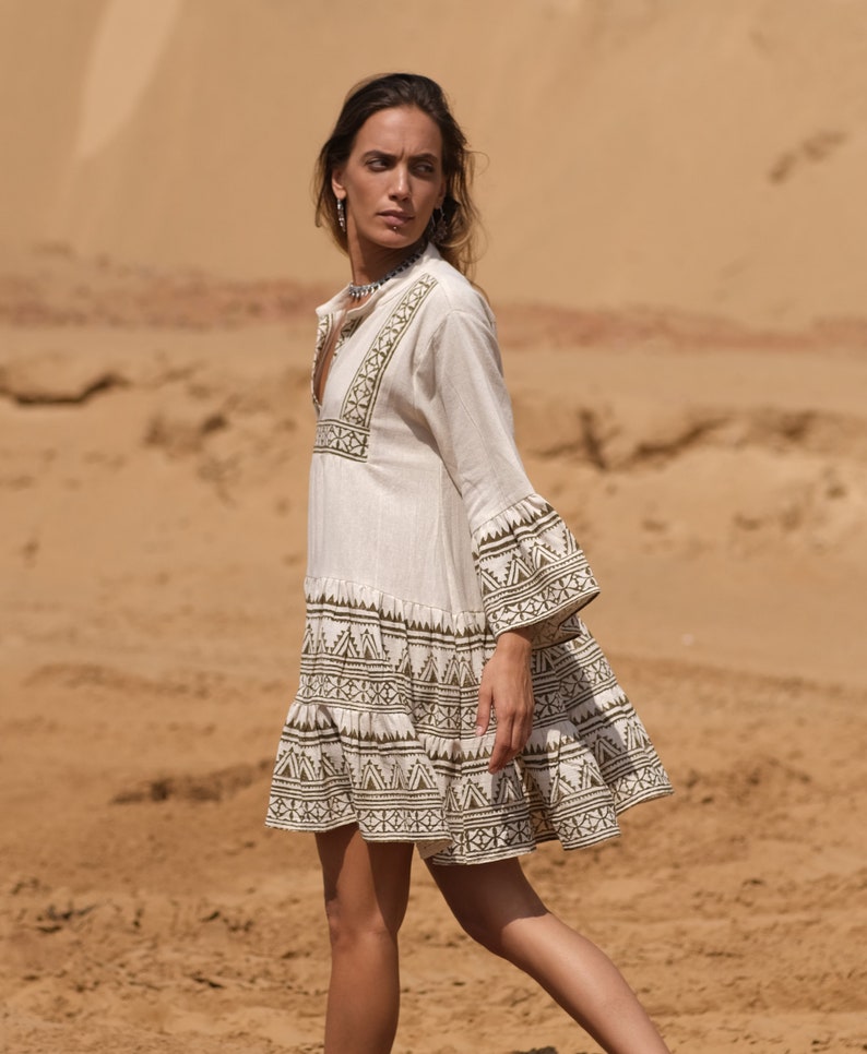 May include: A cream-colored, short dress with long sleeves and a flared skirt. The dress features intricate geometric patterns in a dark olive green color around the neckline, sleeves, and hem. The dress is made of a lightweight fabric, perfect for warm weather.