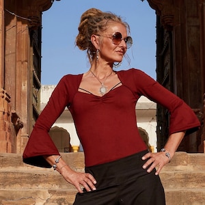 May include: A woman wearing a maroon long-sleeved top with bell sleeves and a scoop neck. She is wearing black pants and has a silver necklace with a pendant. She is also wearing sunglasses and earrings.