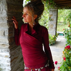 May include: A woman wearing a maroon wrap top with long sleeves and a silver belt. She is standing in front of a stone wall and looking to the right.